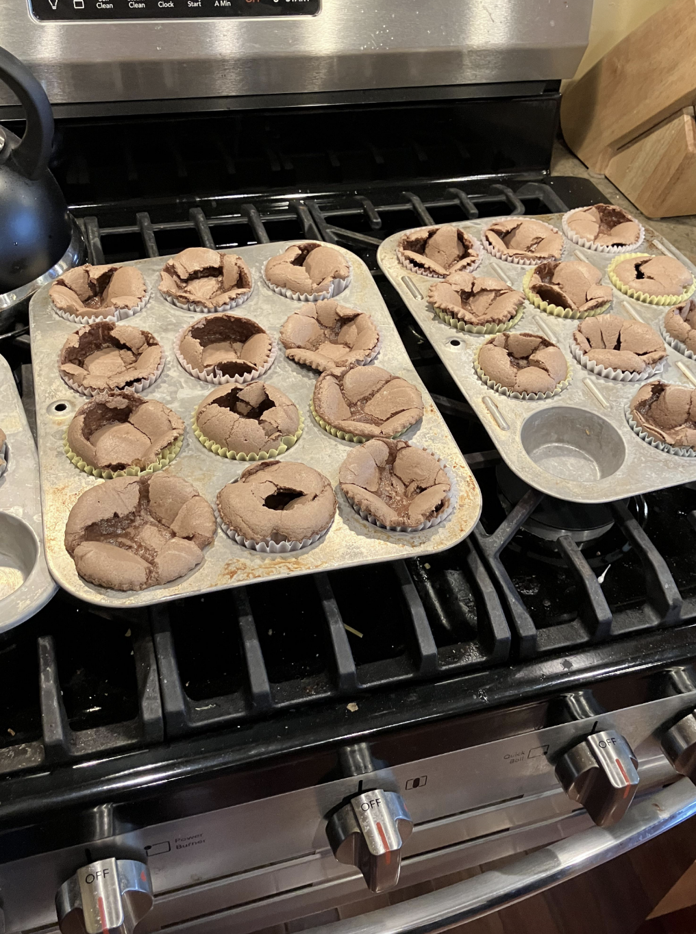 Two muffin trays with several cracked chocolate muffins rest on a stovetop. Some muffin cups are empty