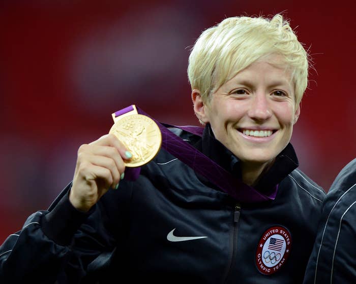 Megan Rapinoe tersenyum sambil memegang medali emas.  Dia mengenakan jaket atletik hitam dengan logo Tim Olimpiade AS