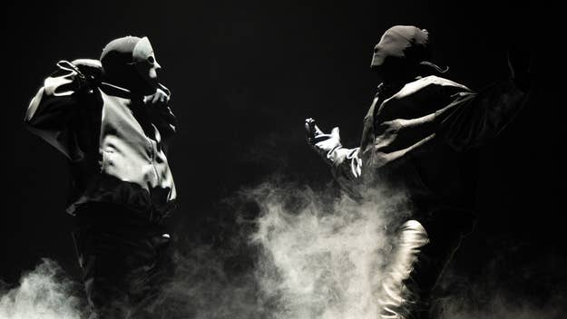 Two masked performers on stage amid smoke, wearing oversized, abstract clothing. Image used in a music-related article. Names of persons unknown