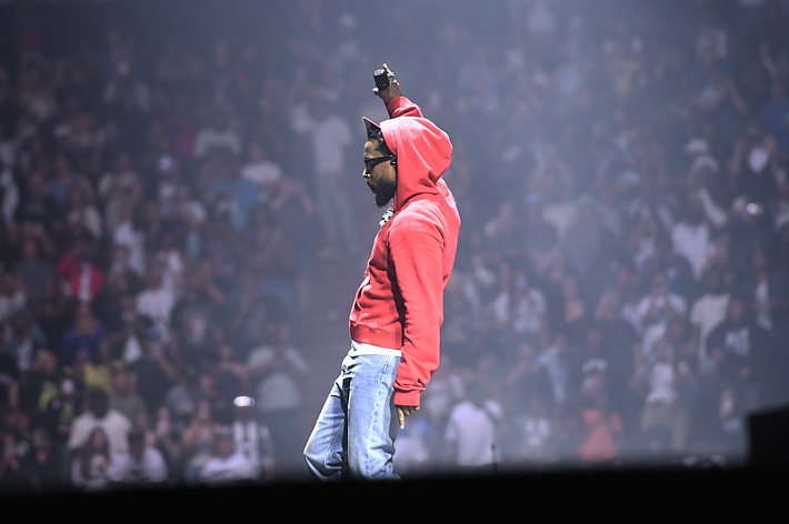 Kendrick Lamar on stage, wearing a hoodie and jeans, raising his right hand with a microphone in front of a large concert audience