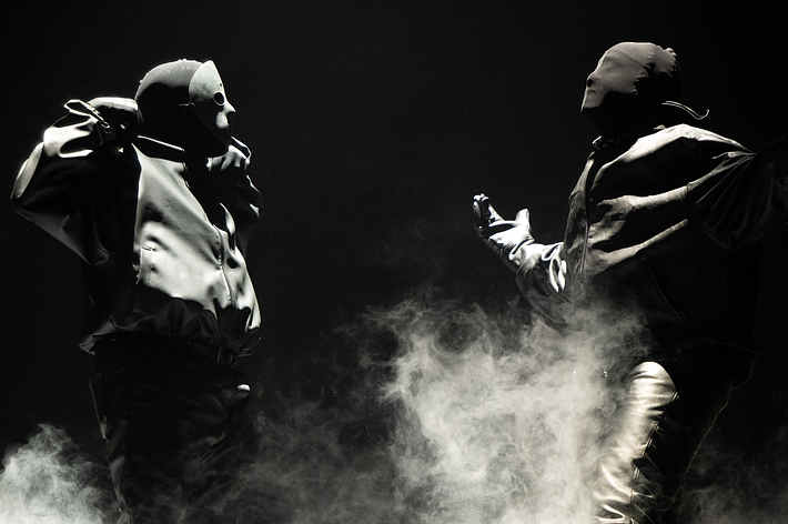 Two masked individuals in heavy jackets and pants perform on a foggy stage