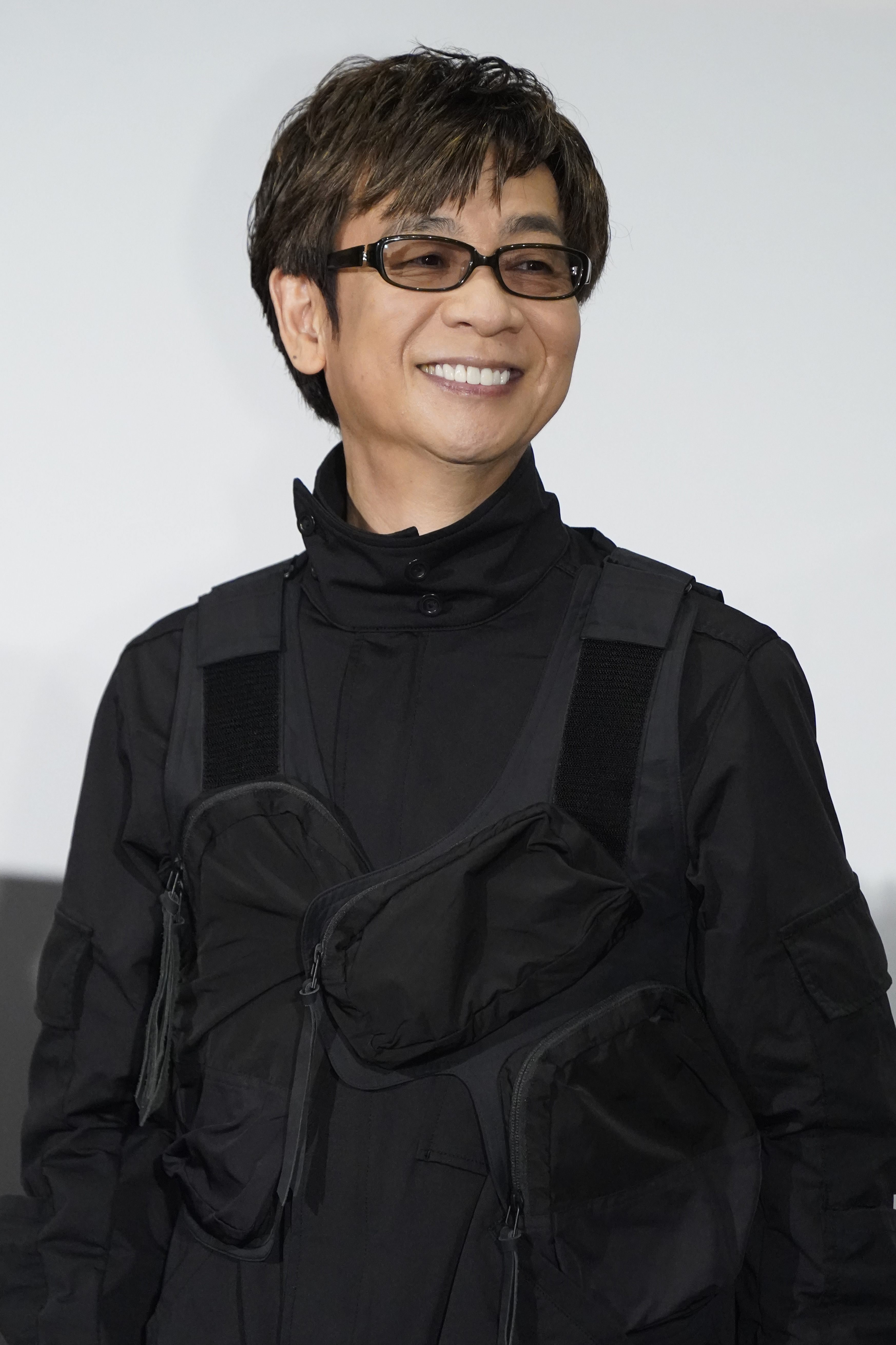 山寺宏一が笑顔で写真に写っている。彼はメガネをかけ、モダンなスタイルの黒い服を着ている。