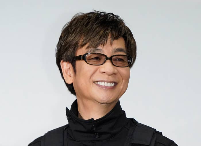 山寺宏一が笑顔で写真に写っている。彼はメガネをかけ、モダンなスタイルの黒い服を着ている。