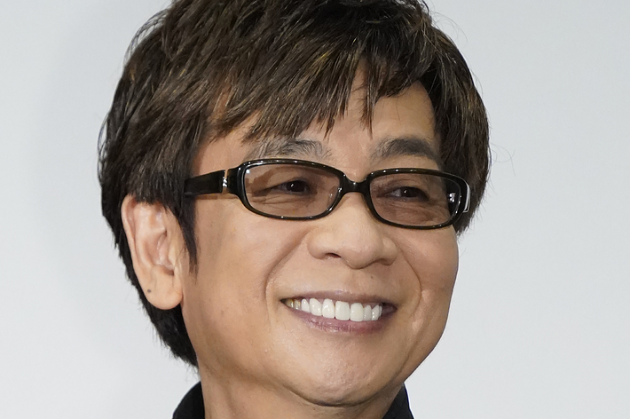 山寺宏一が笑顔で写真に写っている。彼はメガネをかけ、モダンなスタイルの黒い服を着ている。