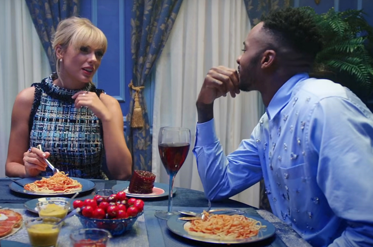 Taylor Swift and a man people enjoy a pasta dinner with drinks and cherries on a table, engaged in conversation, in the Lover music video