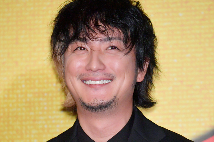 満島ひかりが黒のスーツにネクタイを着て笑顔でポーズしています。