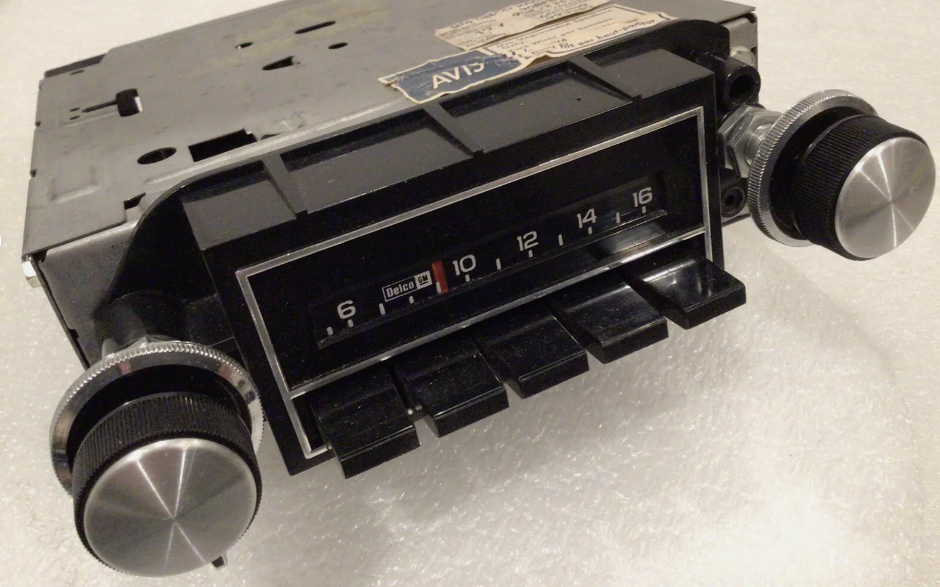 Vintage car radio with black knobs, a tuning dial, numbered scale from 6 to 16, and labeled "Delco." The unit has visible wear and a partial sticker