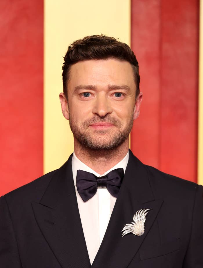 Justin Timberlake wearing a suit with a bow tie and a brooch, standing in front of a red carpet backdrop