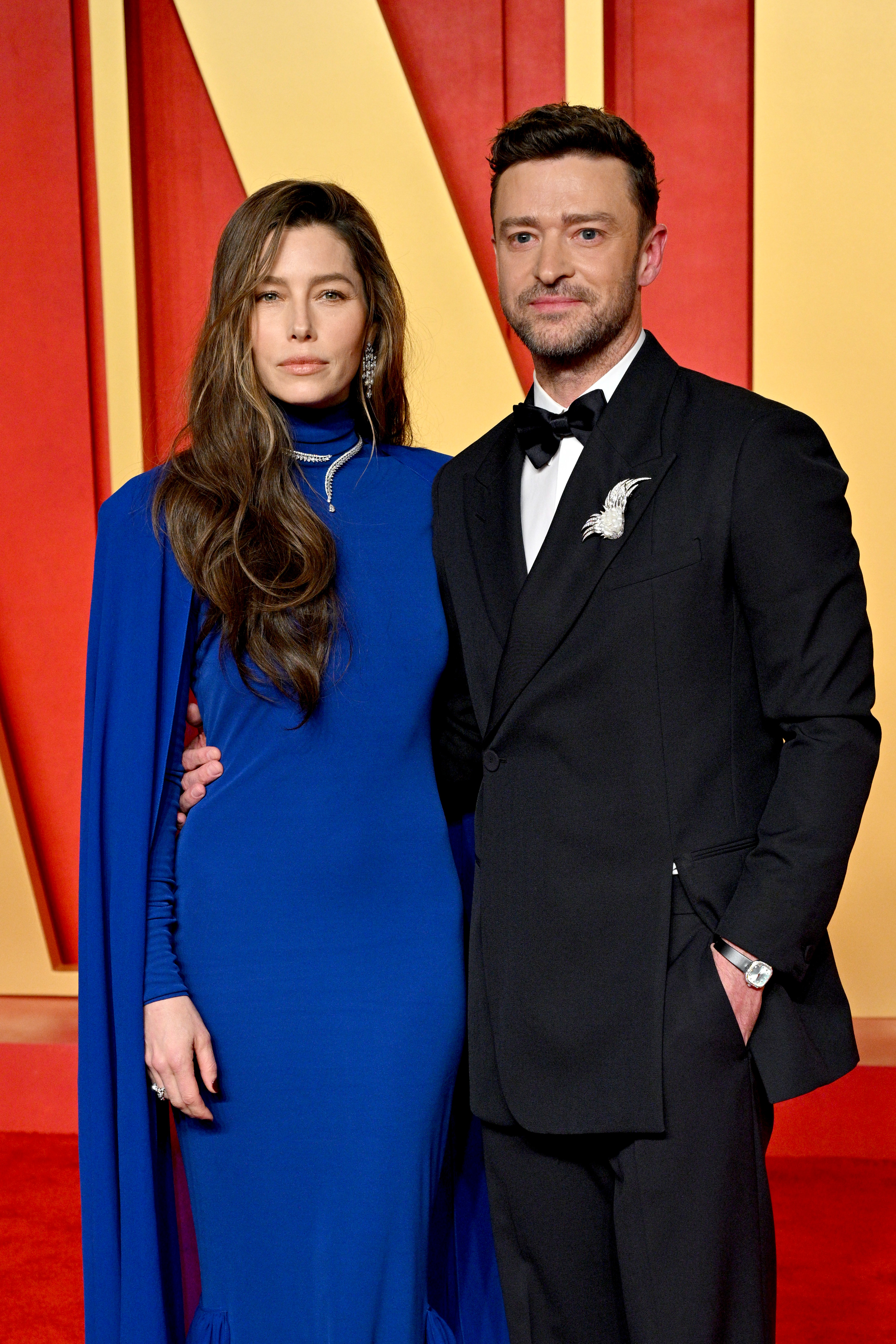 Jessica Biel and Justin Timberlake on the red carpet. Jessica wears a long, elegant dress with a cape accent, and Justin is in a classic tuxedo with a bow tie