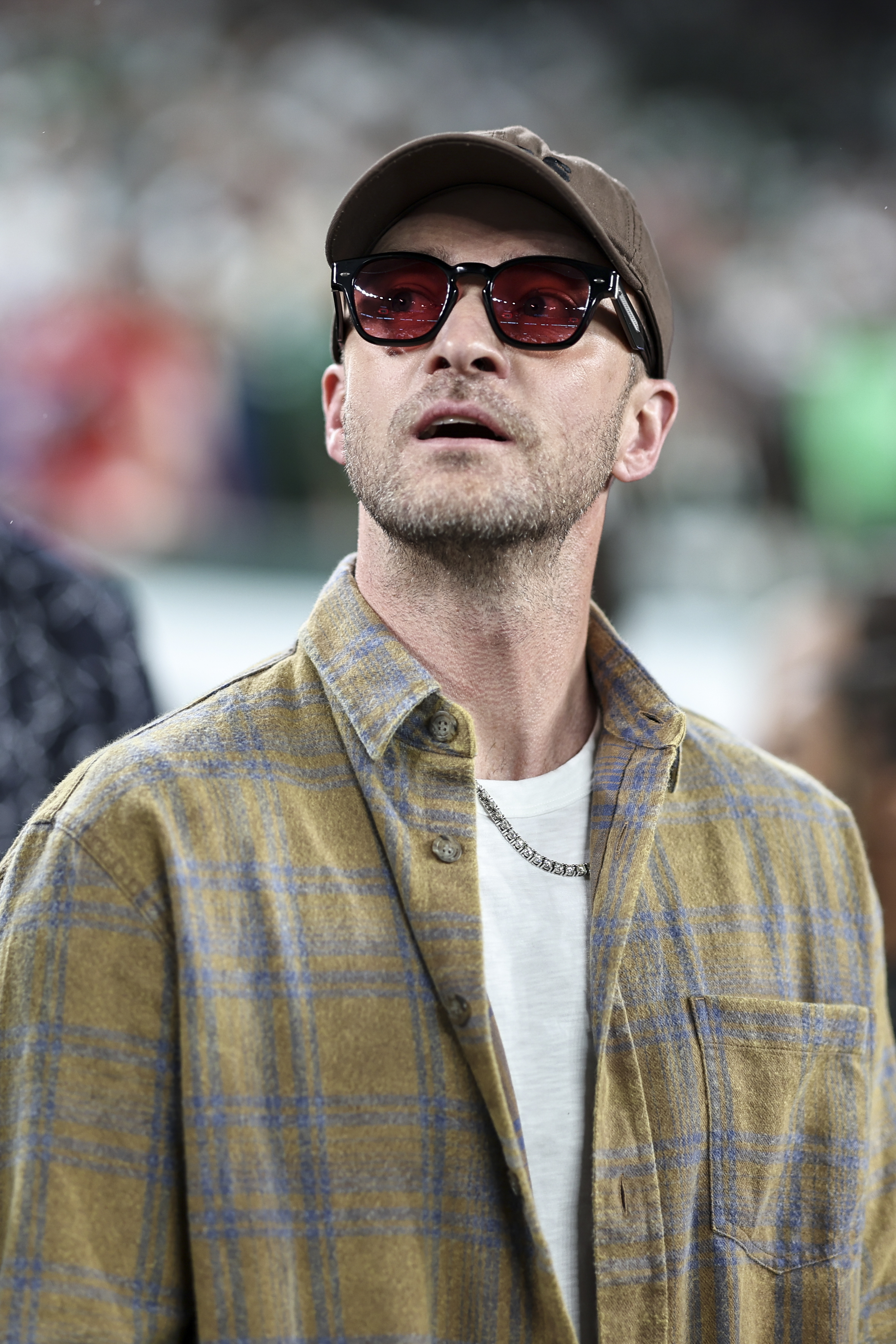 Justin Timberlake wears a plaid shirt, t-shirt, chain necklace, tinted sunglasses, and a cap. He looks upward with a blurry background behind him