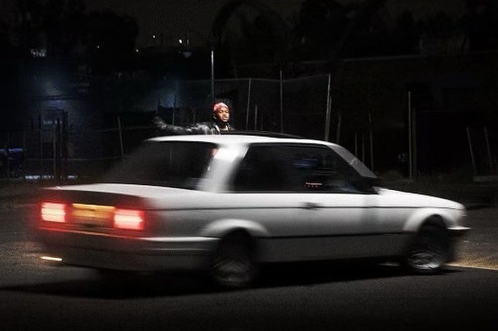 A person is standing through the sunroof of a moving car at night in an urban area. The image has a "Parental Advisory: Explicit Content" label in the corner