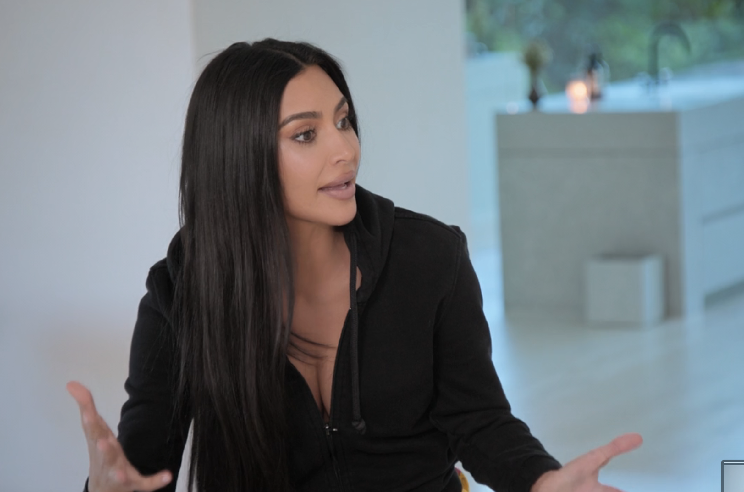 Kim Kardashian sits at a table, gesturing with her hands, in a modern, minimalist kitchen