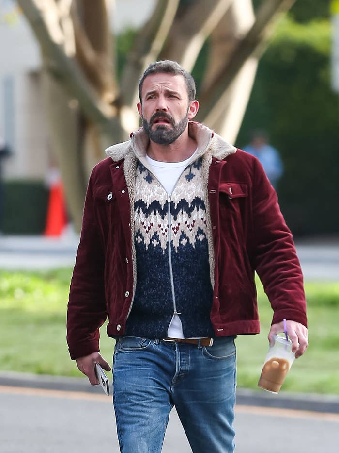 Ben Affleck walks outside wearing a jacket over a patterned sweater, holding an iced coffee and a smartphone