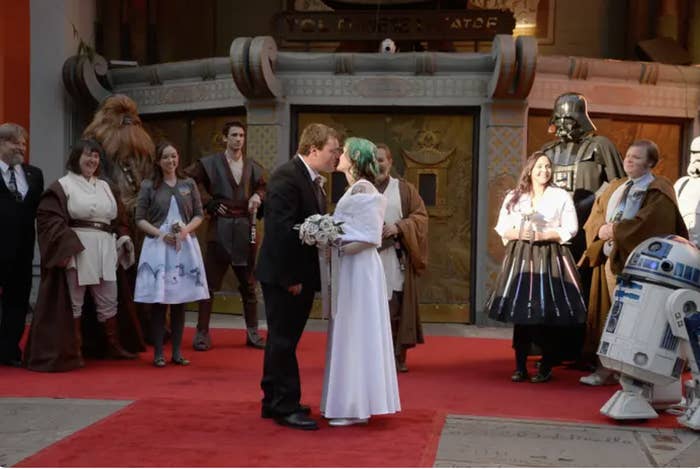 A couple dressed as Star Wars characters kiss at their wedding. Characters like Darth Vader, Chewbacca, R2-D2, and others stand around them on a red carpet