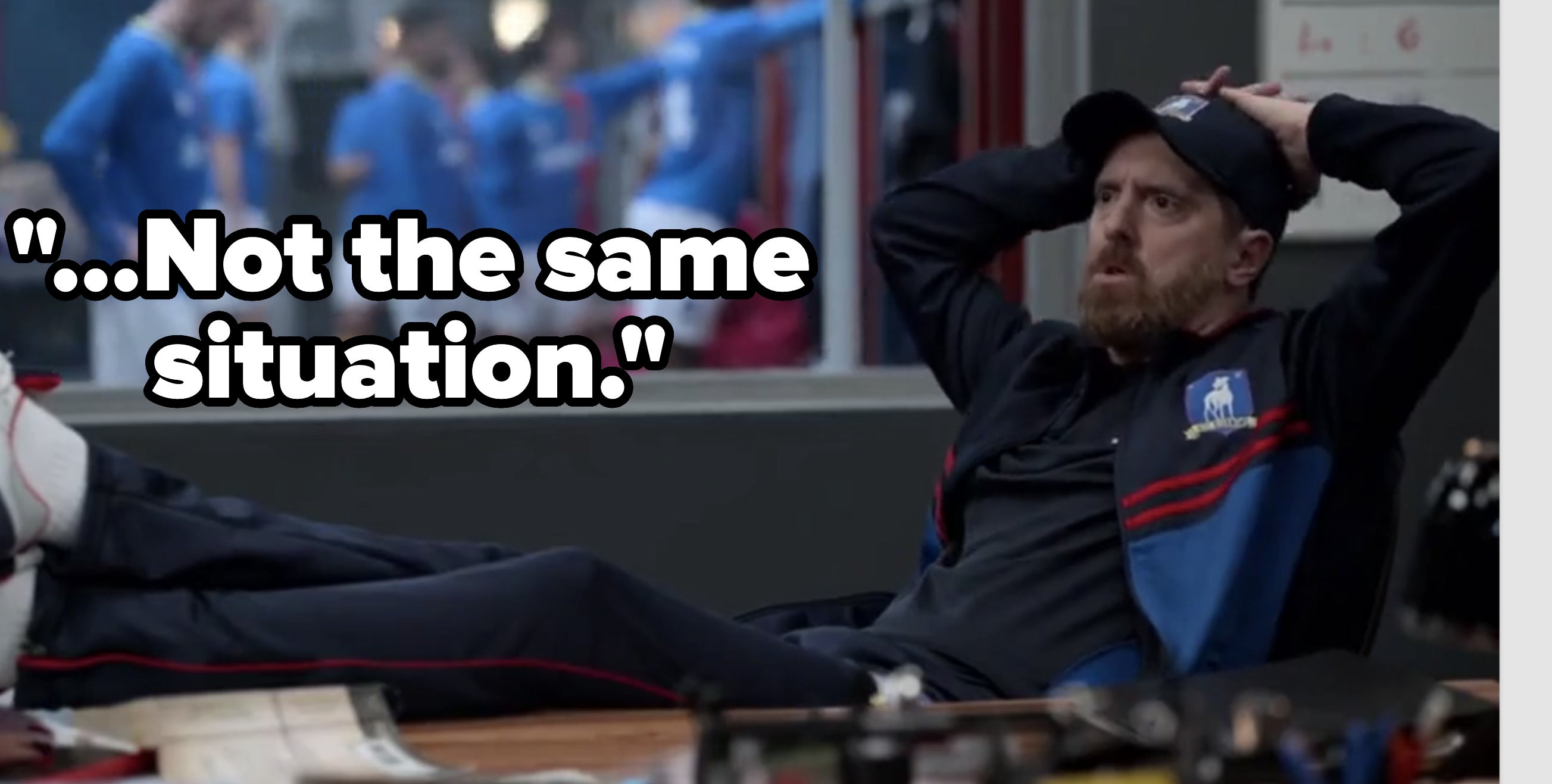 Bearded man in sportswear sits with legs on desk, hands on head. Soccer team in blue jerseys visible in the background