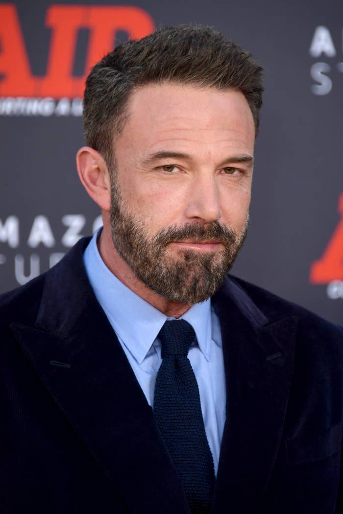 Ben Affleck on the red carpet, wearing a dark suit jacket, light-colored shirt, and a dark tie