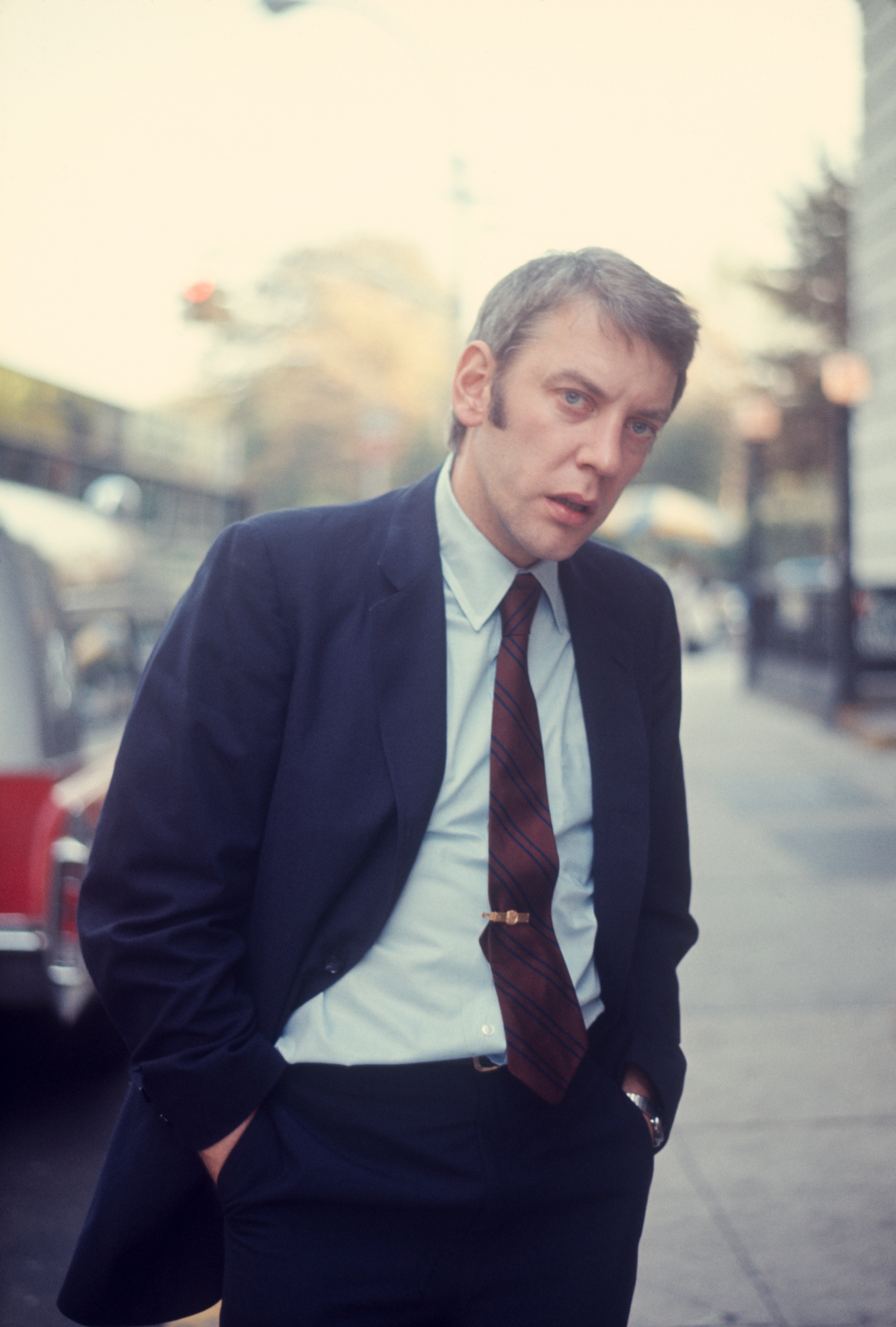 Donald Sutherland in a suit, standing on a street with hands in his pockets, looking pensive