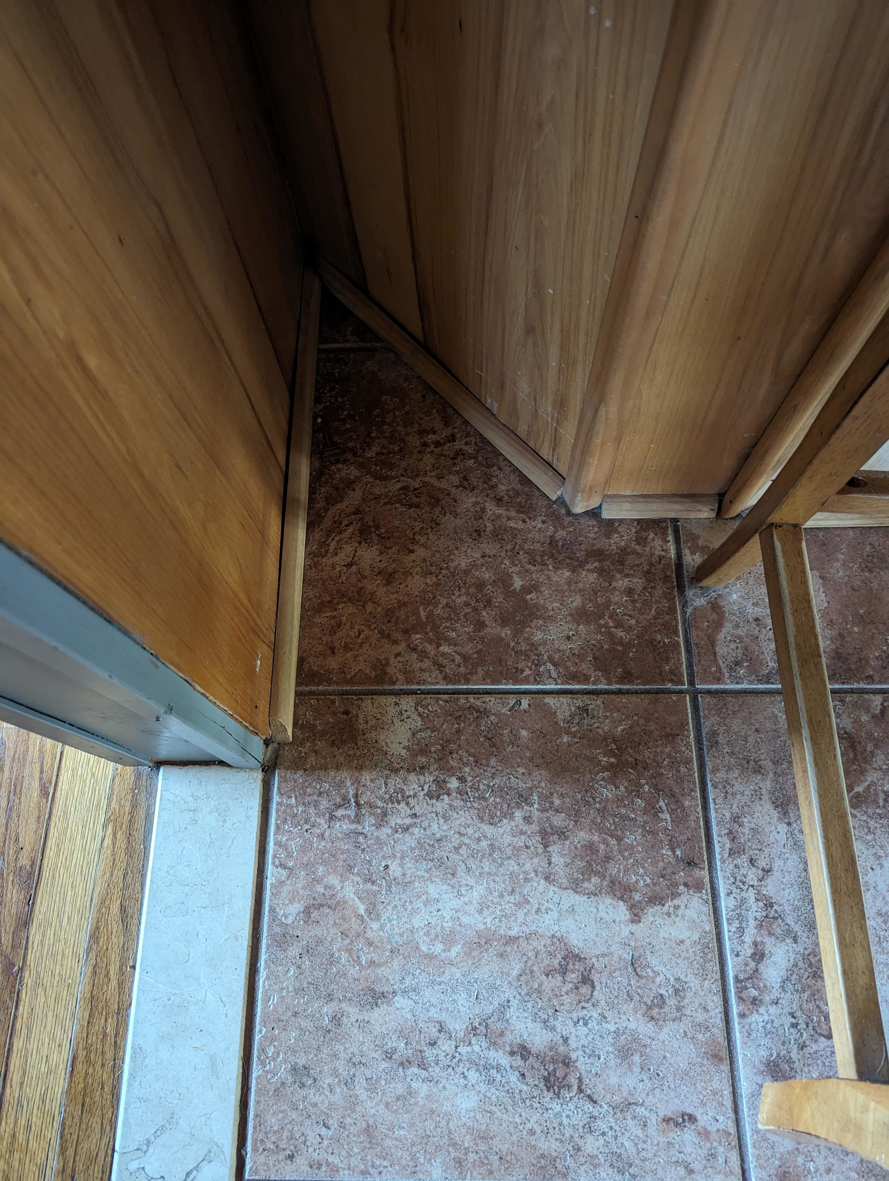 Visible corner of a room with tiled and wooden floors. Misaligned tiles and wood trim forming a narrow, awkward triangular gap