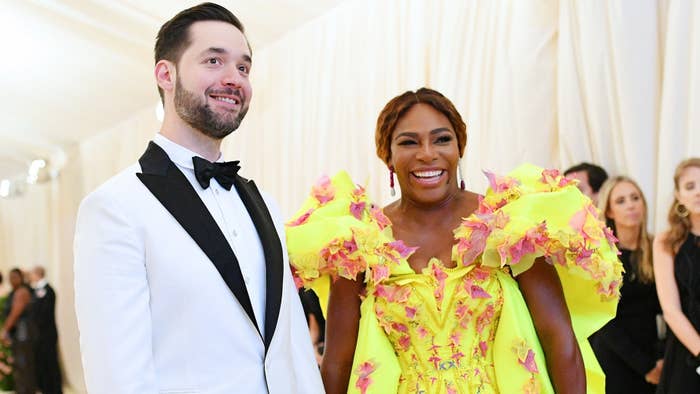 Alexis Ohanian and Serena Williams on the red carpet. Alexis in a white tuxedo, Serena in a bright dress with voluminous floral embellishments. Both smiling