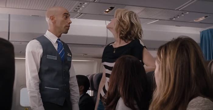 A flight attendant speaks to a woman standing in the airplane aisle while other passengers are seated and watching
