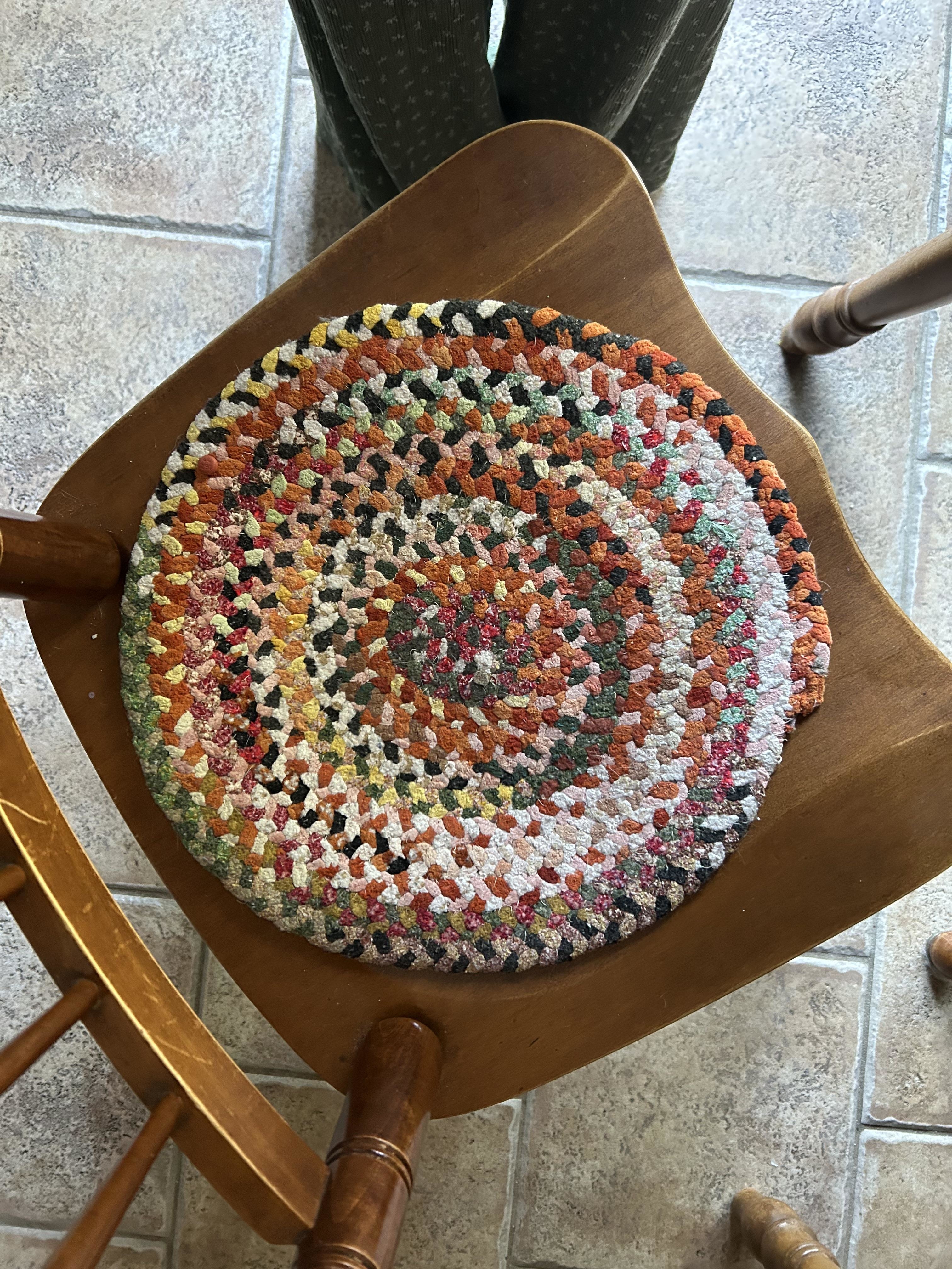 Crocheted multicolor round cushion on a wooden chair, viewed from above. A person&#x27;s legs are partially visible in the background, standing on a tiled floor