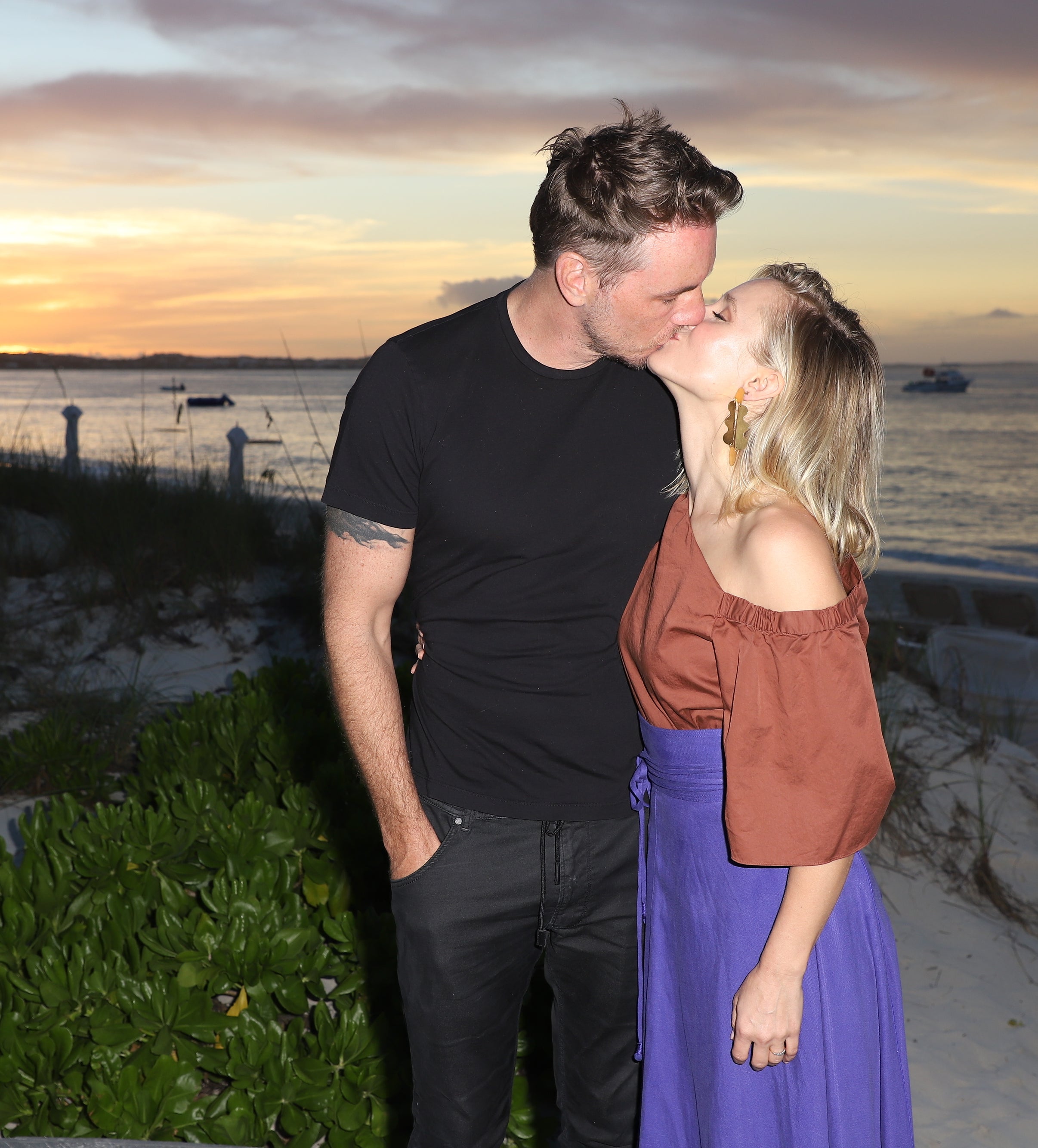 Dax Shepard and Kristen Bell kissing on a beach at sunset, wearing casual attire