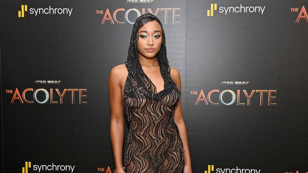 Amandla Stenberg on "The Acolyte" red carpet in a sheer, patterned dress with a hood, in front of a background displaying "The Acolyte" and Synchrony logos