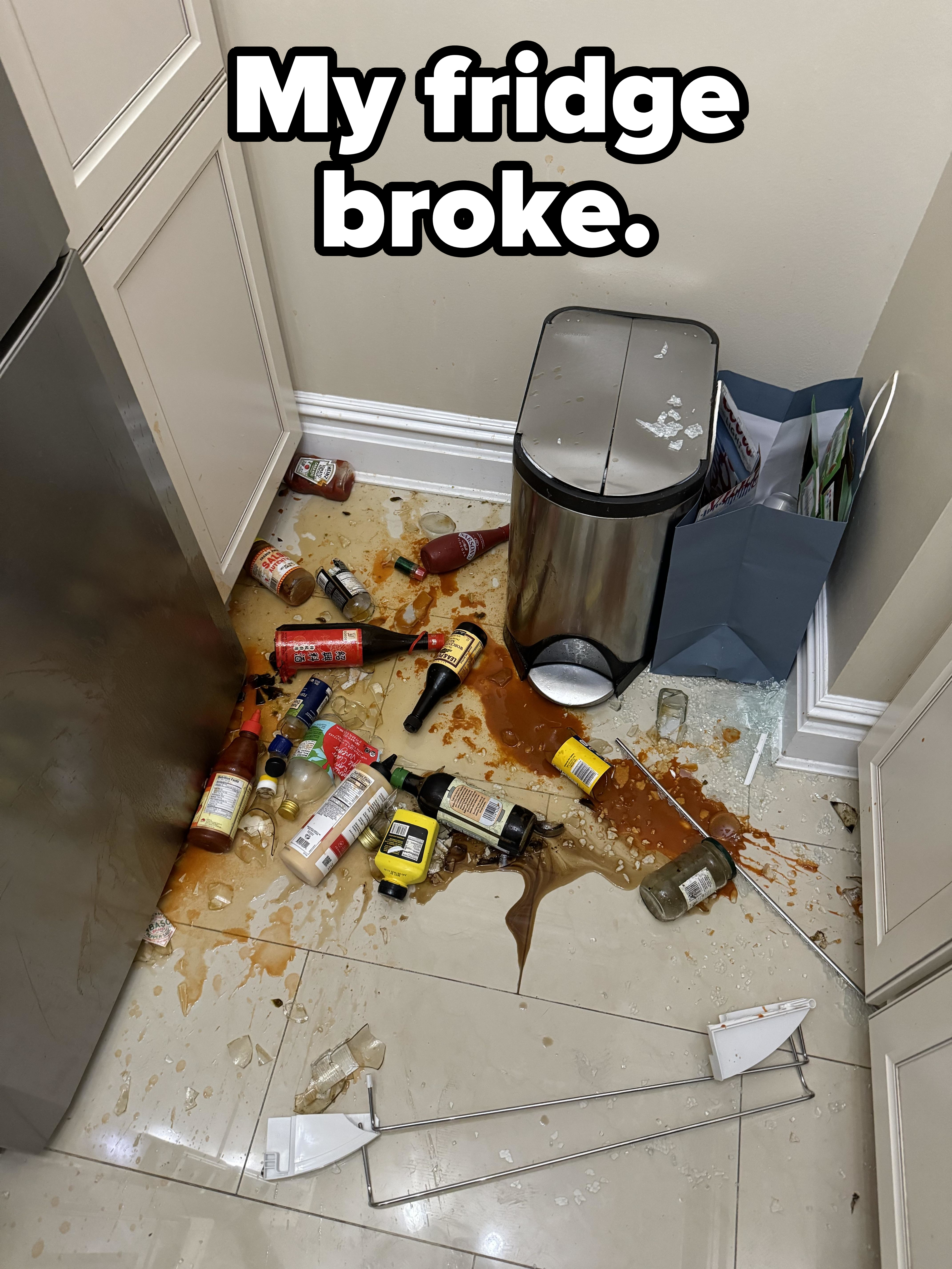 Kitchen corner with a tipped-over trash can spilling bottles and liquid on the floor, creating a mess. Broken glass and scattered recycling items are also visible