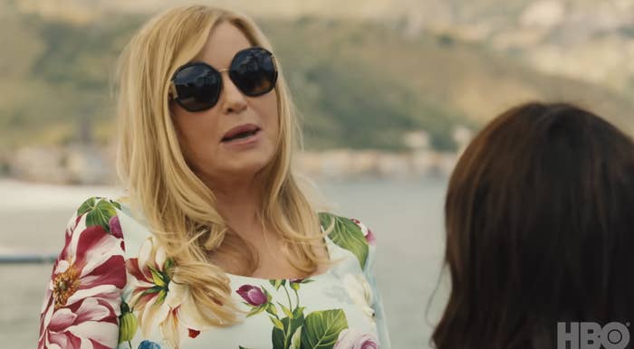 Jennifer Coolidge, wearing black sunglasses and a floral dress, interacts with another person against a scenic backdrop