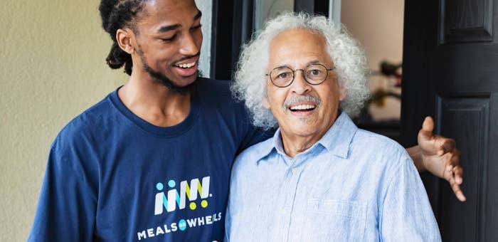 A young man in a "Meals on Wheels" shirt stands smiling arm in arm with an older man in a button-down shirt at a front door