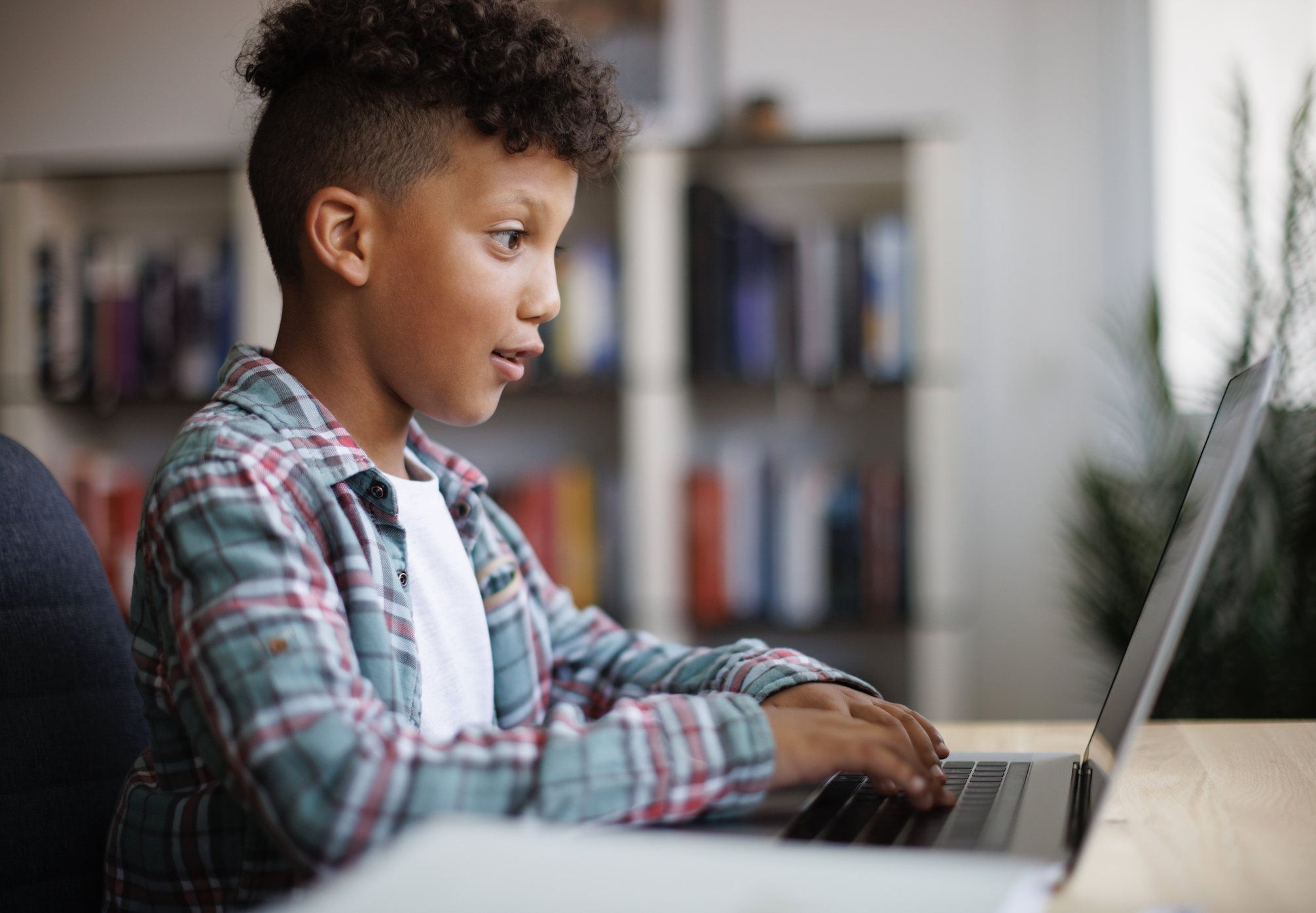 A young boy in a plaid shirt and t-shirt typing on a laptop focused on the screen while sitting. Bookshelves are in the background
