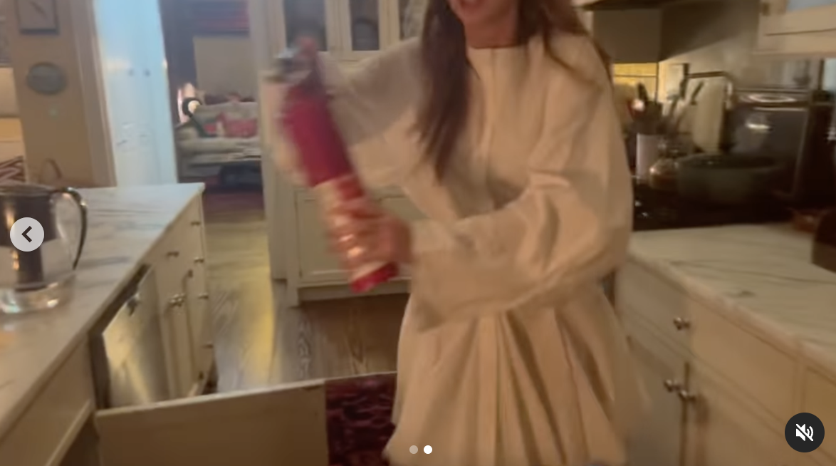 A woman in a white blouse holds a red fire extinguisher in a kitchen