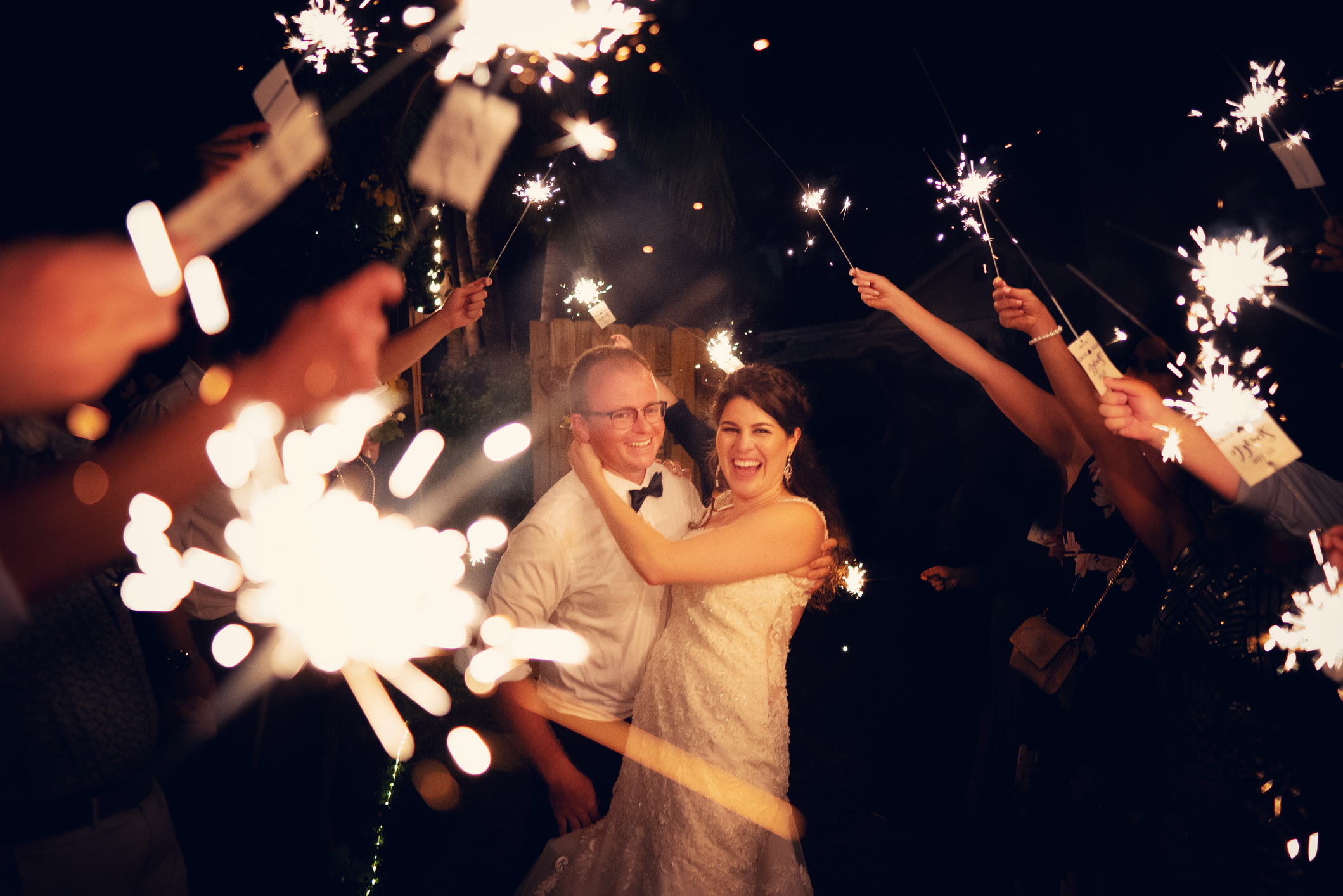 A joyful bride in a wedding dress and groom in a bow tie are surrounded by guests holding sparklers during the night