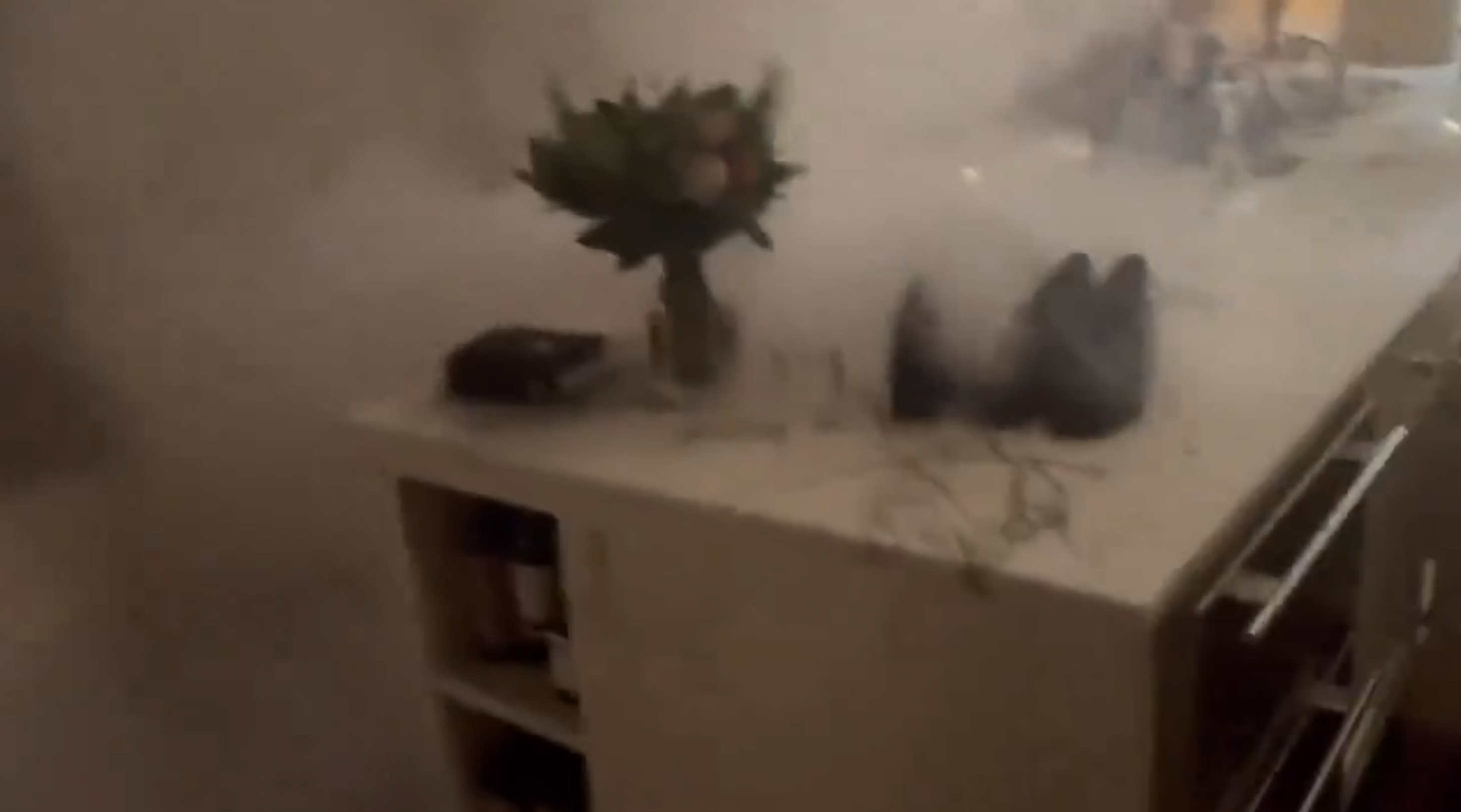 A kitchen island with flowers and shoes on it is surrounded by smoke or mist, with kitchen items and a shelf visible in the background