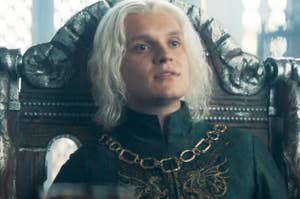 Tom Glynn-Carney as Aegon II Targaryen sitting on a throne, wearing a dark, regal outfit with ornate details, in a scene from a TV show