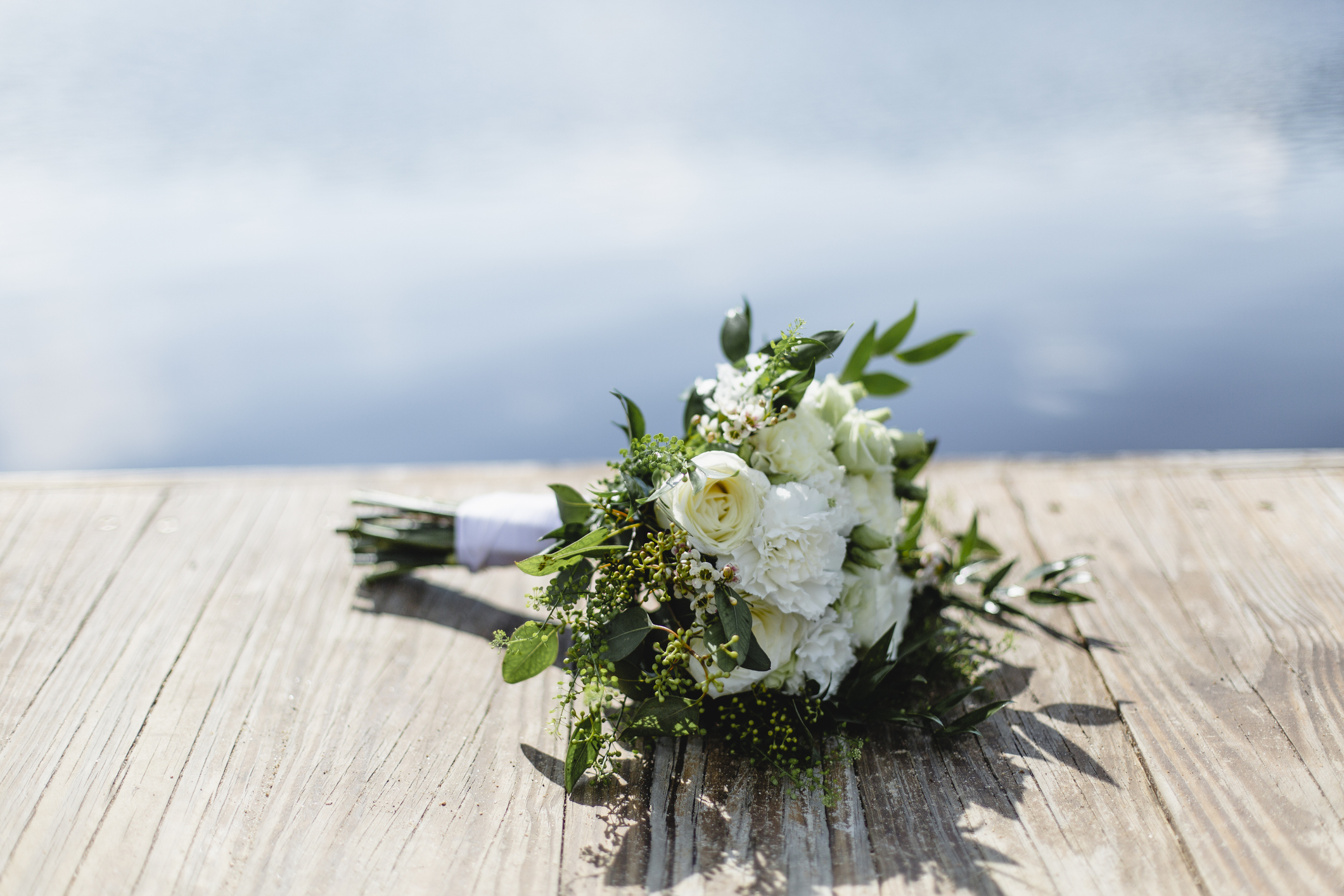 A close-up of a white and green wedding bouquet lies on a wooden dock by the water