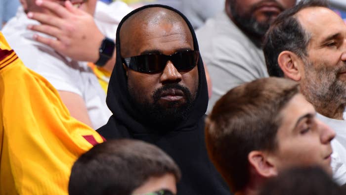 Ye wearing a black hoodie and sunglasses, sitting among a crowd at an event