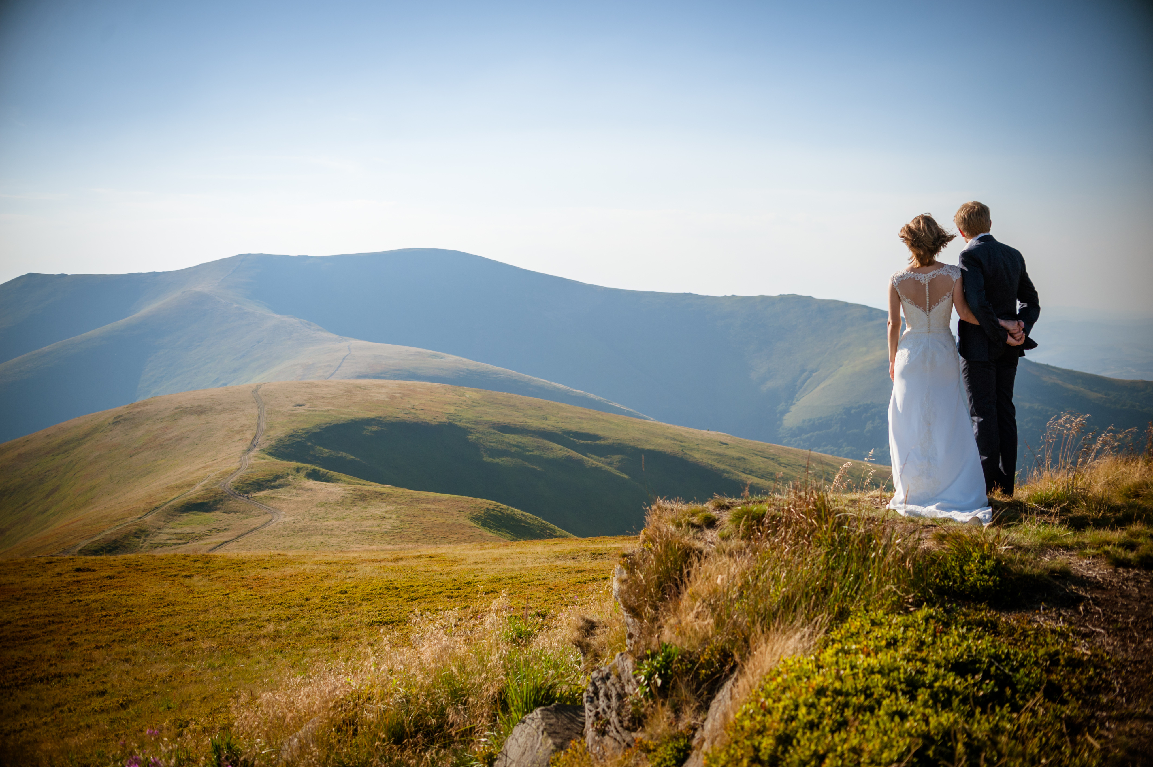 A couple in wedding attire stands on a grassy hilltop overlooking distant mountains and valleys, facing away from the camera