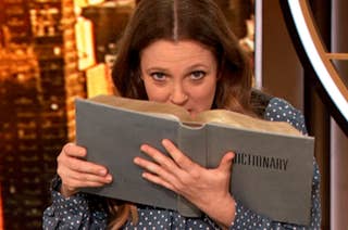 Drew Barrymore playfully peeks over a large dictionary while wearing a polka dot blouse