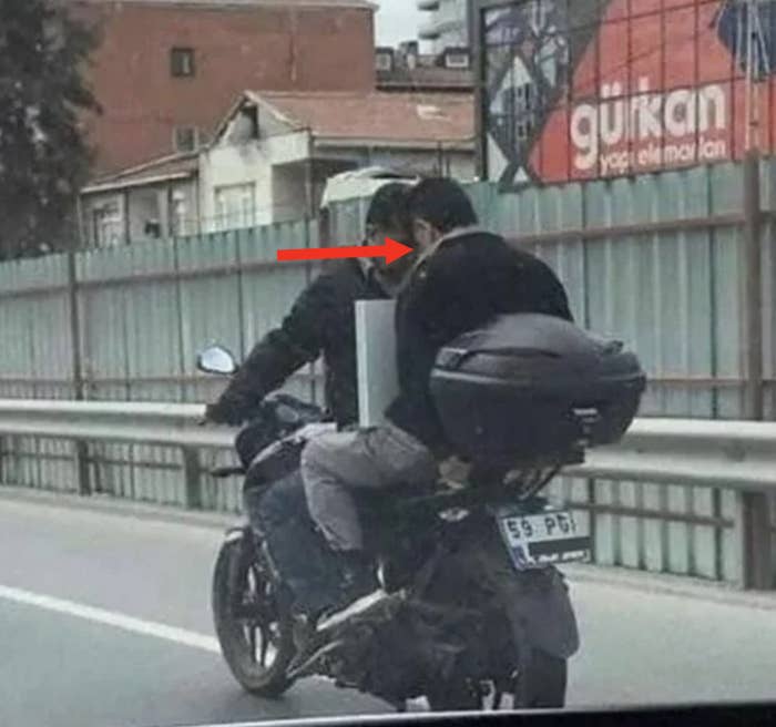 Two men on motorcycles parked side by side on the road, talking to each other. One motorcycle has a large rear case; both riders wear protective gear