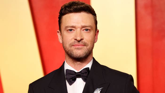 Justin Timberlake in a formal suit with a bow tie, standing on a red carpet background