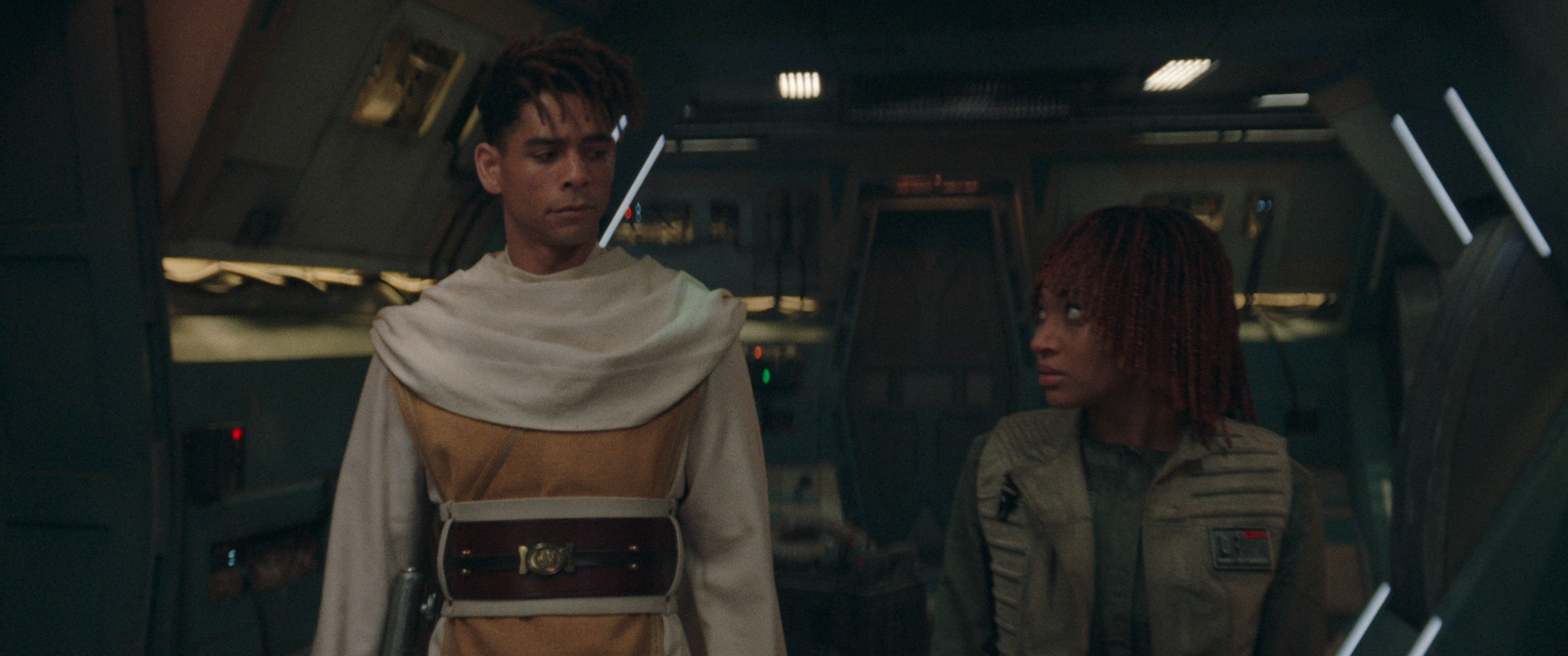Jay Lycurgo and Ellora Torchia in a futuristic spaceship set, Jay in a robe with a belt, and Ellora in a tactical outfit looking at each other