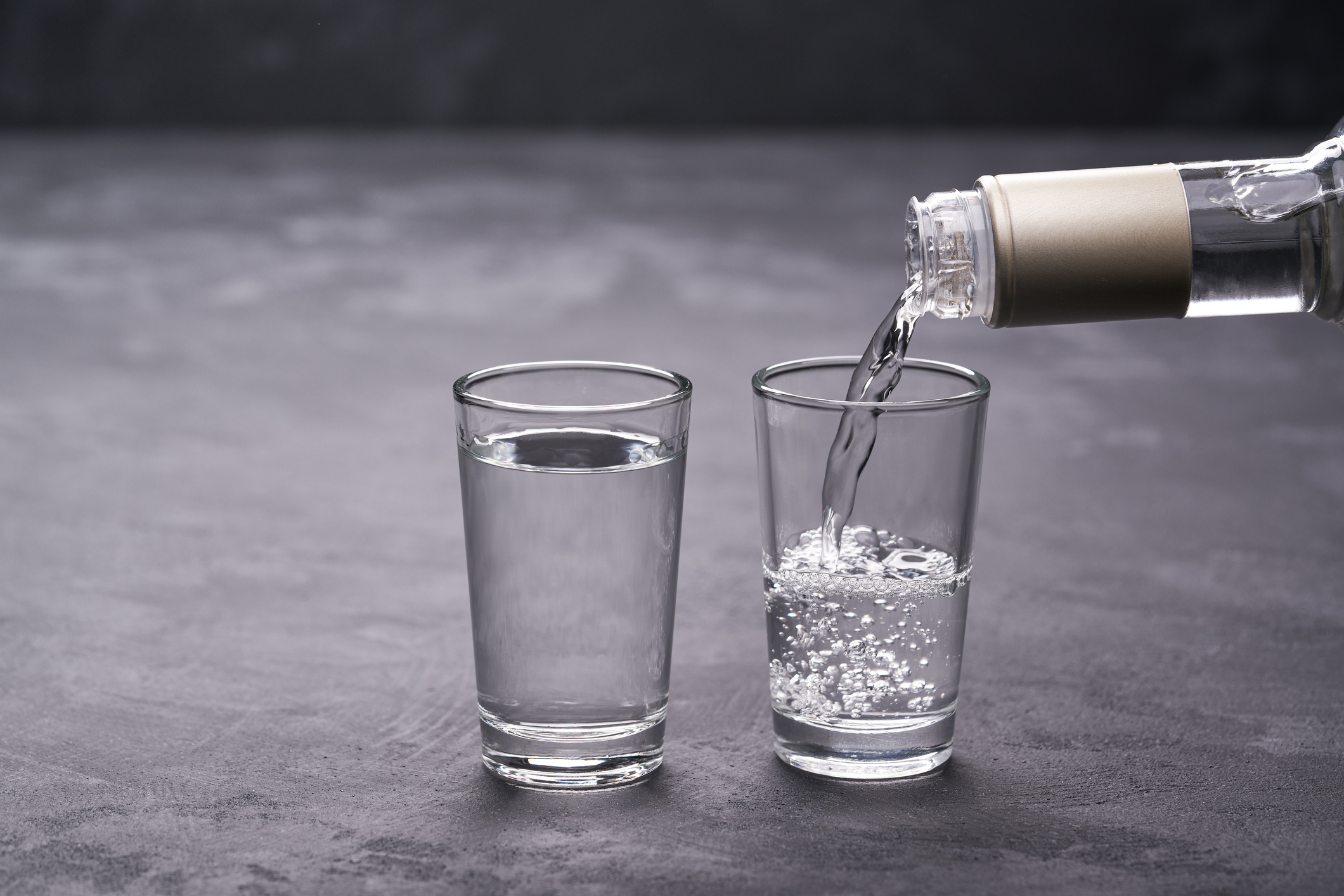 A bottle is pouring clear liquid into a glass, positioned beside another glass filled with clear liquid