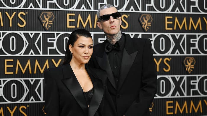 Kourtney Kardashian and Travis Barker on the red carpet, dressed in matching black suits, attending an event with FOX and Emmys logos in the background