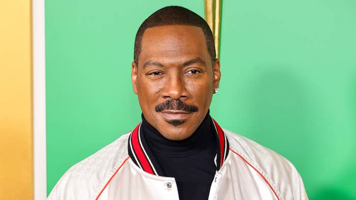 Eddie Murphy wearing a stylish white jacket with a black turtleneck, standing in front of a colorful background