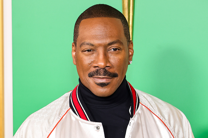 Eddie Murphy wearing a stylish white jacket with a black turtleneck, standing in front of a colorful background