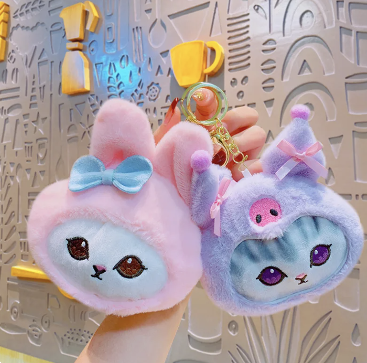 Mano sosteniendo dos llaveros de peluche con forma de gatos, uno rosa con lazo azul y otro morado con lazo rosa, sobre fondo decorativo