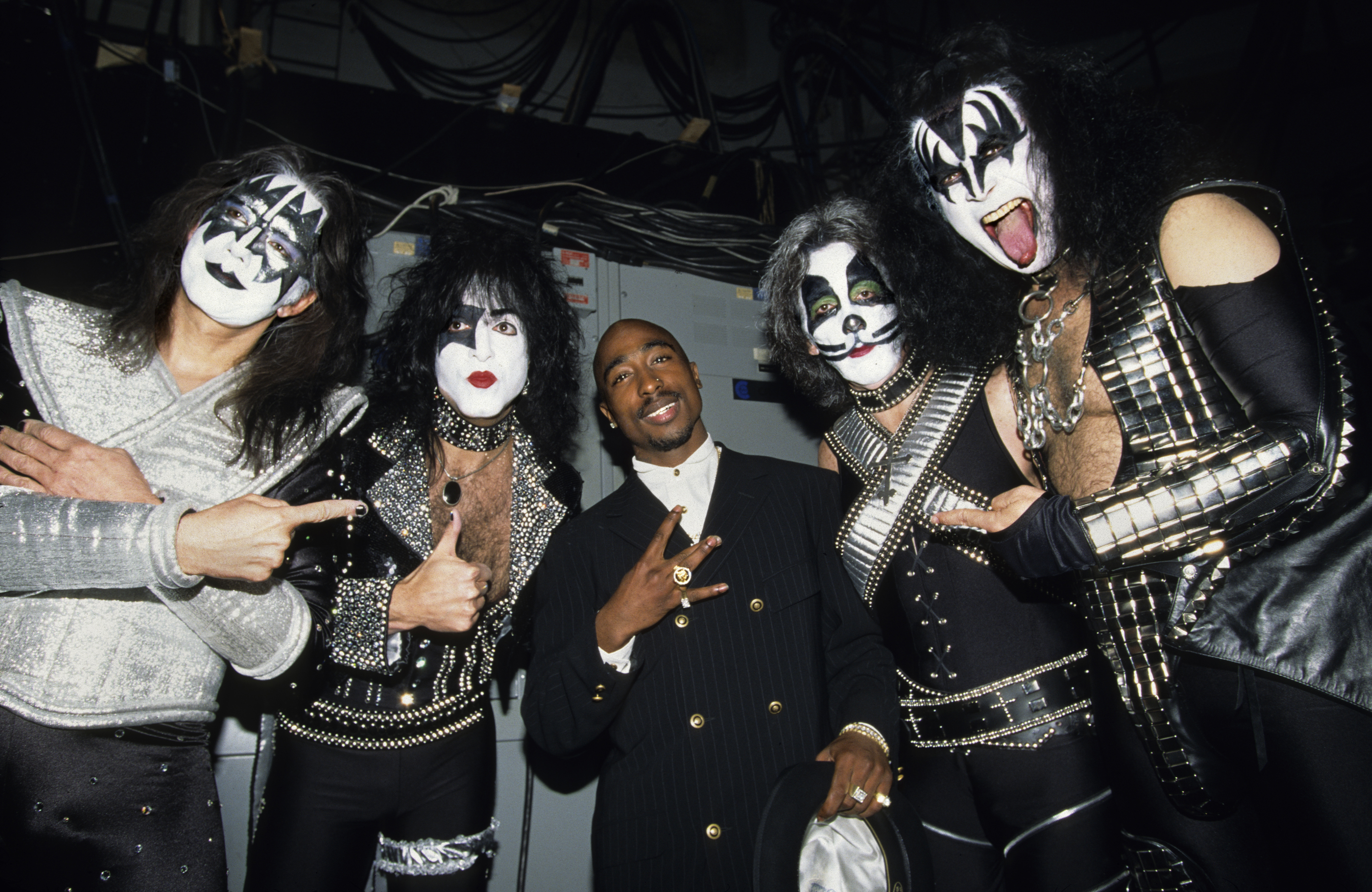 Four members of the rock band KISS, in full stage makeup and costumes, pose with Tupac Shakur, who is wearing a suit and holding a white item