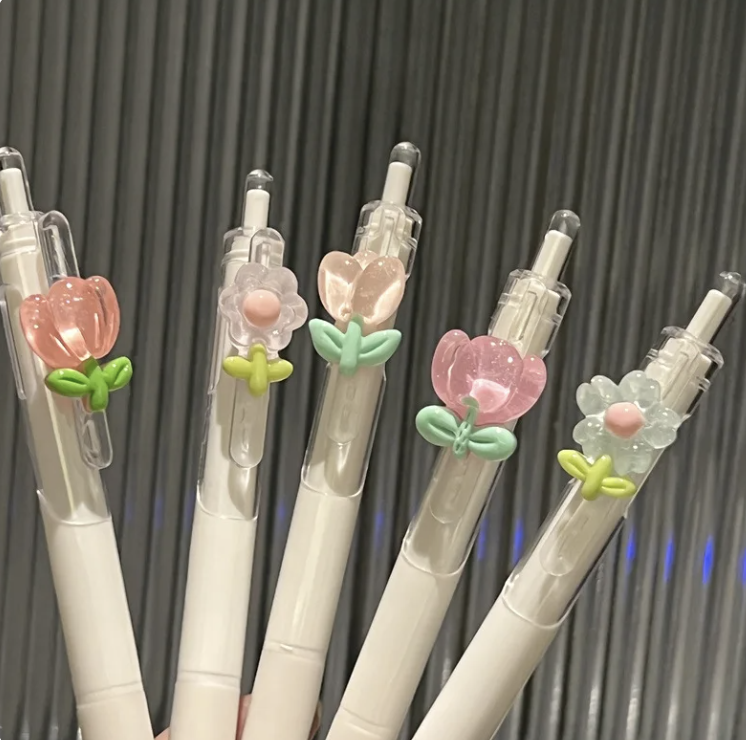 Cinco bolígrafos blancos con adornos de flores y corazones en diferentes diseños en la parte superior de cada bolígrafo