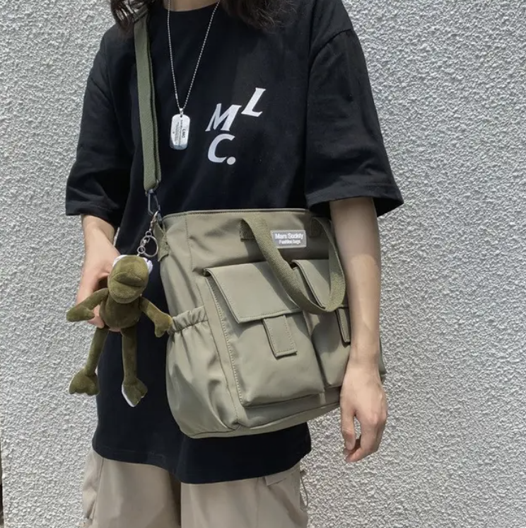 Persona con camiseta negra, pantalones beige y bolsa verde con un llavero de rana